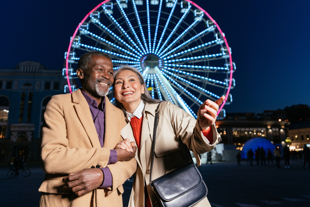 "A multiethnic senior couple smiling and enjoying their evening in front of a brightly lit Ferris wheel at night