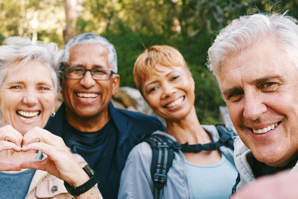 Group of four senior friends smiling during a nature walk, with one woman making a heart shape with her hands and lush greenery in the background.