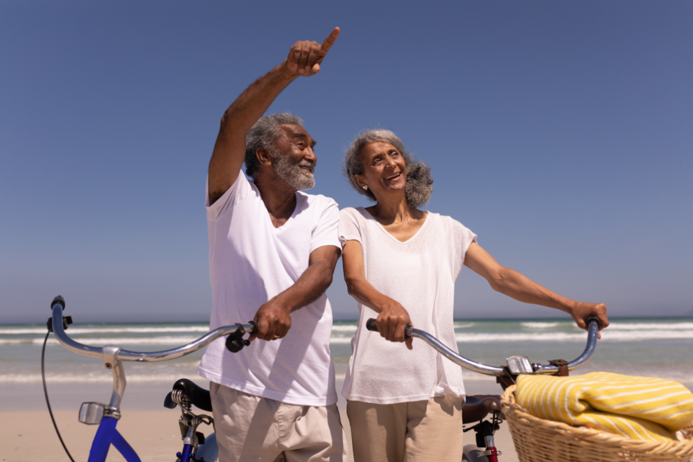 An older couple standing with bicycles on a beach, the man pointing towards the sky, both smiling and enjoying the sunny day.