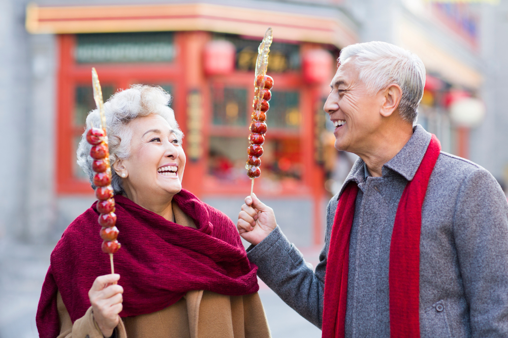 Elderly couple holding candied fruit sticks, smiling at each other, dressed warmly in matching red scarves, with colorful street food stalls in the background.