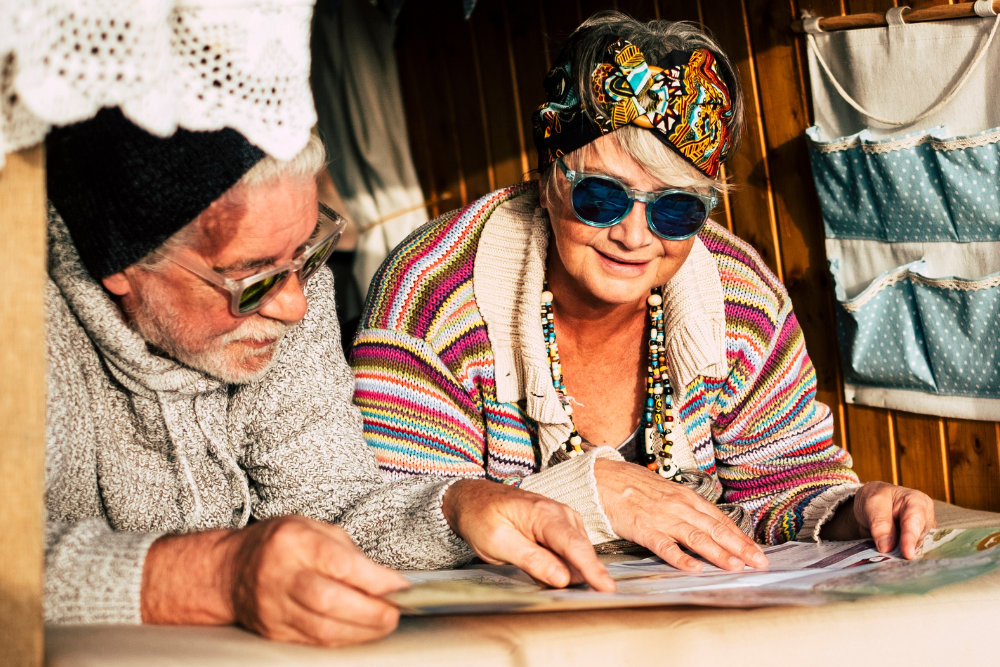 An older couple wearing colorful clothes and sunglasses, looking at a map together, enjoying their time in a cozy, decorated space.