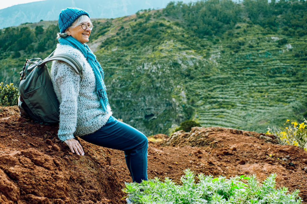 An older woman wearing a blue beanie, glasses, and a cozy sweater, sitting on a hillside with a backpack, enjoying a scenic view of green terraces and mountains.