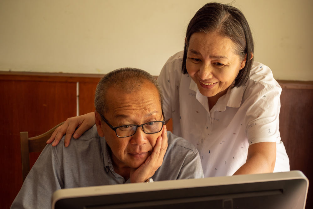 Senior couple using a computer together, with the man focused on the screen and the woman smiling supportively.