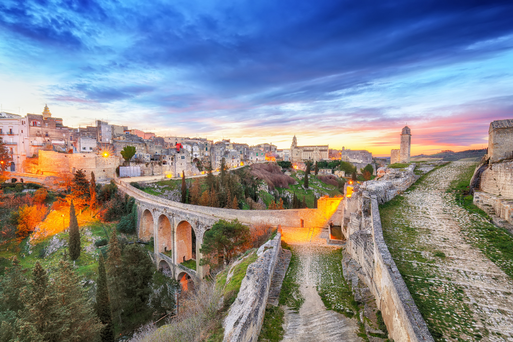 View of the ancient town of Gravina in Puglia, Italy, with a historic bridge and canyon illuminated by warm lights at twilight.