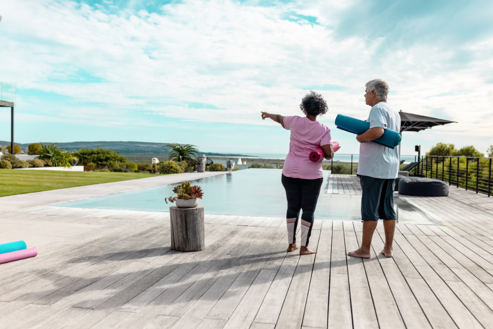 A senior couple standing by a pool with yoga mats, looking at the scenery.