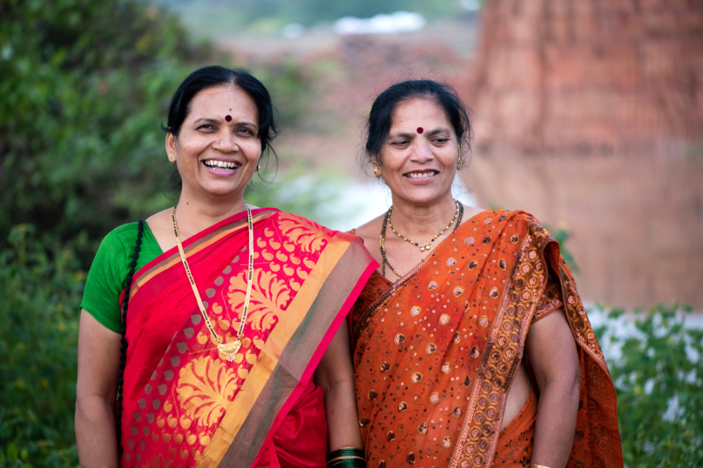 Two women wearing colorful saris, smiling and standing outdoors in a natural setting.