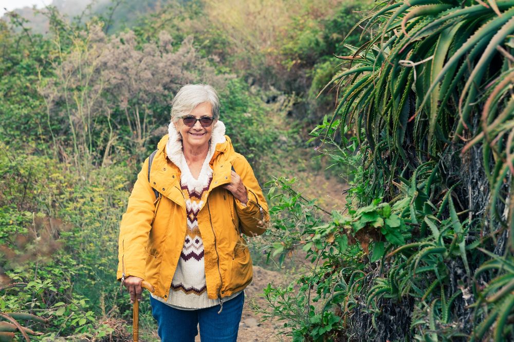 Senior woman in a yellow jacket and glasses hiking on a green trail with a walking stick.