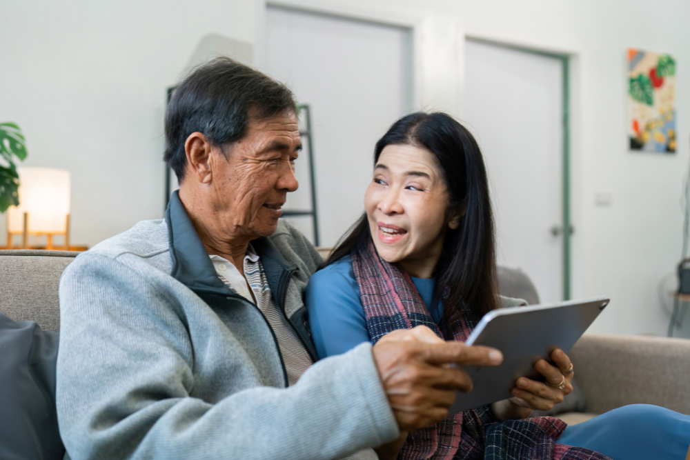 Senior couple sitting on a couch using a tablet together, with the man holding the tablet and the woman smiling warmly at him.