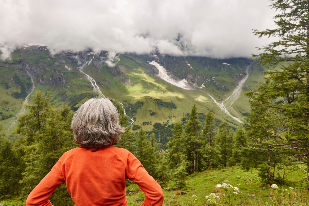 Senior woman with grey hair standing at a mountain viewpoint, looking at a lush green valley with waterfalls and cloudy sky.