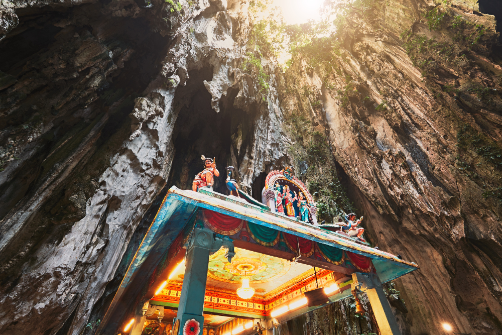 Colorful statues atop a temple structure inside the Batu Caves in Kuala Lumpur, with the cavernous walls and sunlight filtering in from above