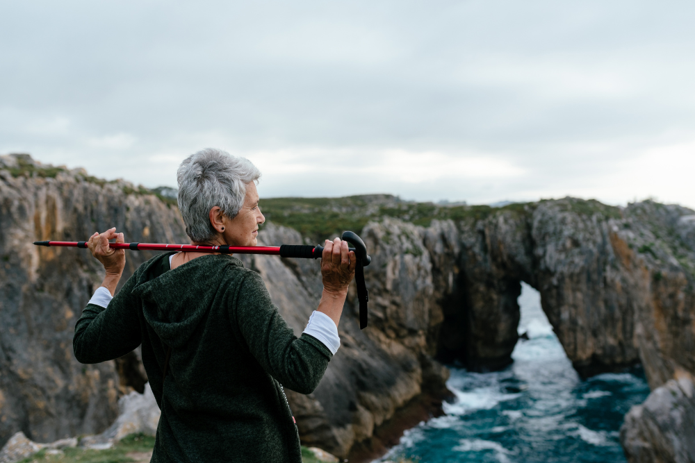 Elderly woman holding a walking stick, enjoying a scenic view of rocky cliffs and the ocean