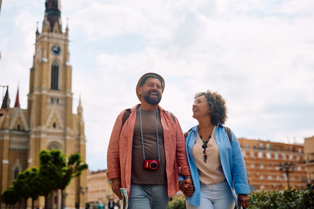 Smiling elderly couple holding hands and walking through a city with a historic clock tower in the background.