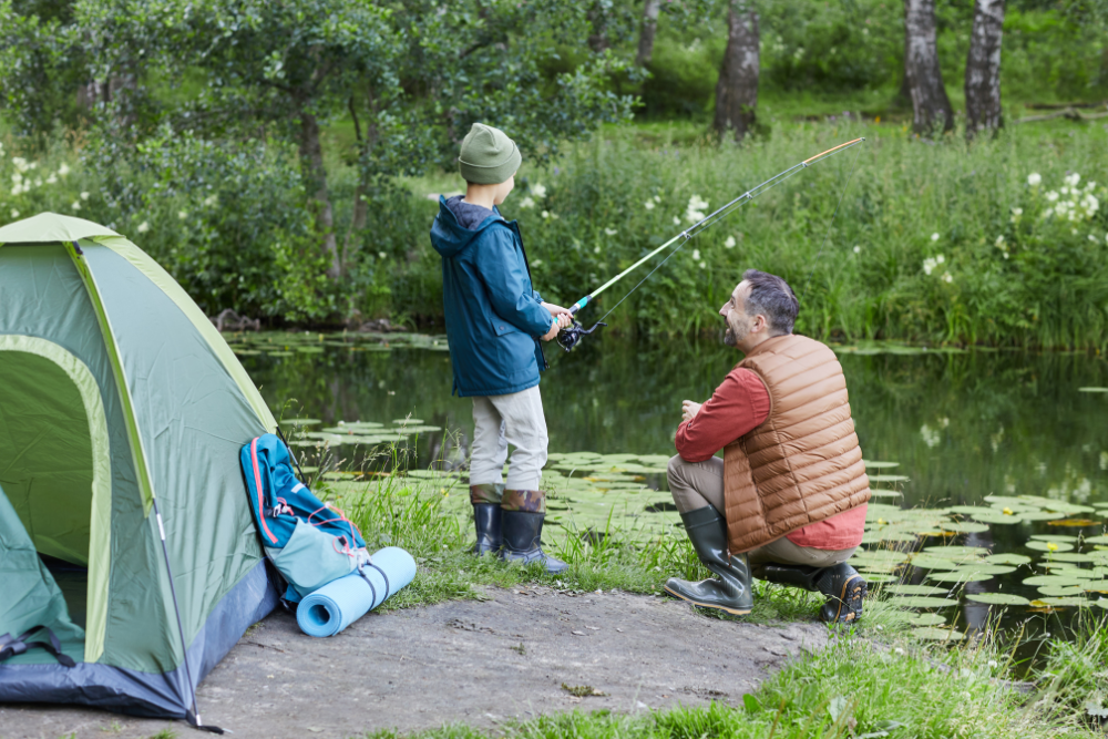 Father and son fishing together by a lake near a campsite.