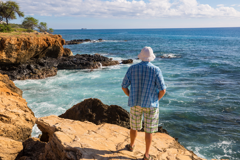 Man wearing a sun hat and casual clothes standing on rocky cliffs overlooking the ocean."