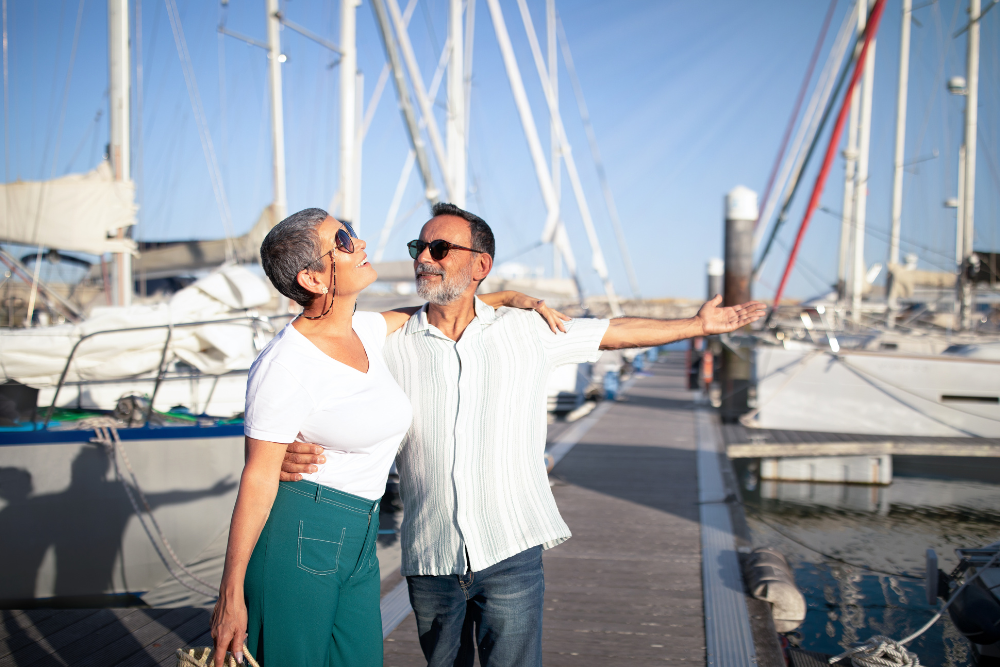 Senior couple enjoying a sunny day at a marina with sailboats in the background.