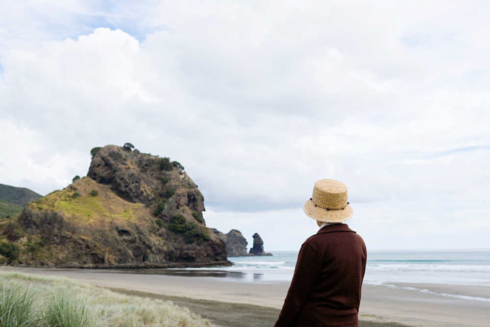 Senior person standing at a beach, looking at a rocky hill formation by the sea.