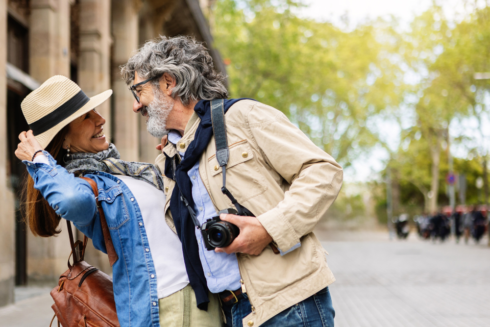 Happy older couple enjoying a day out in a city, with the man holding a camera and the woman wearing a hat and smiling.