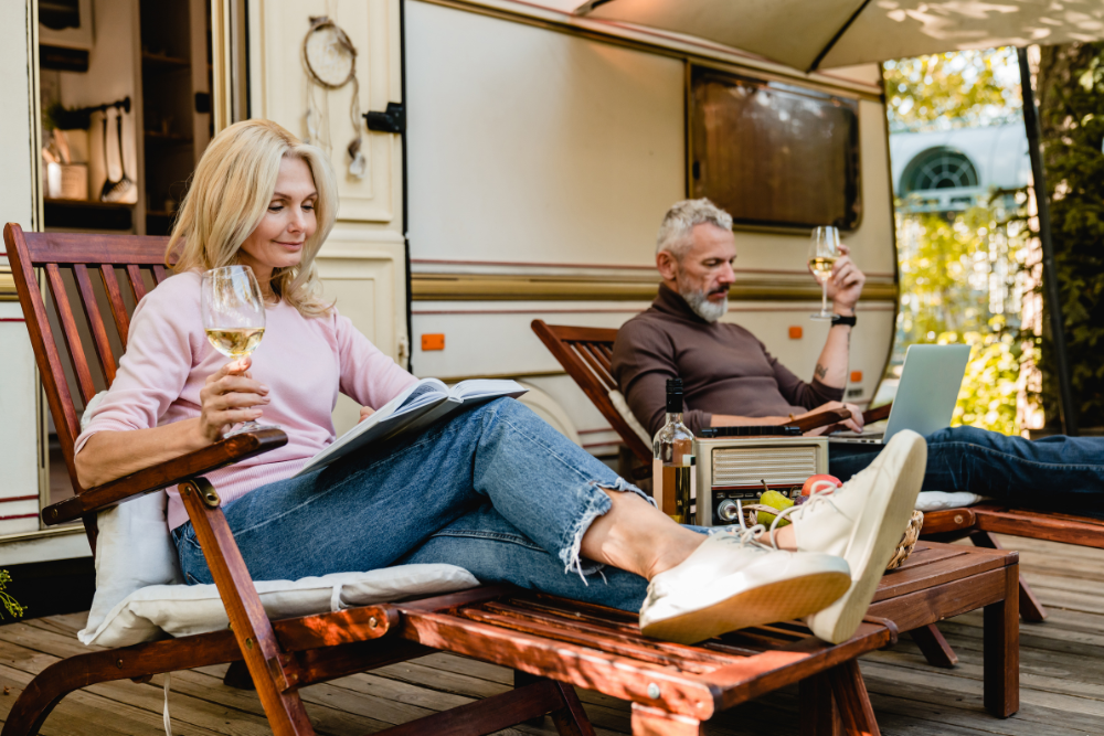 A senior couple relaxing outside their RV. The woman is reading a book and holding a glass of white wine, while the man is using a laptop and also holding a glass of wine. They appear to be enjoying a peaceful, leisurely moment together.