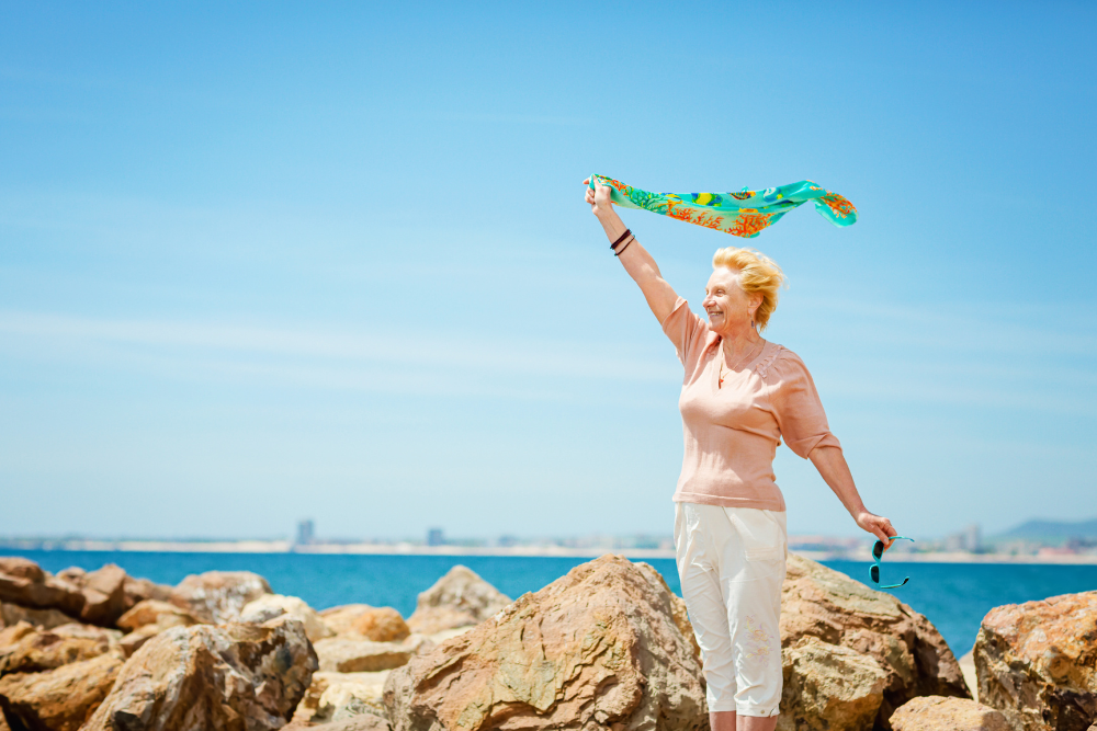 A joyful elderly woman standing on rocks by the sea, holding a colorful scarf above her head on a sunny day.