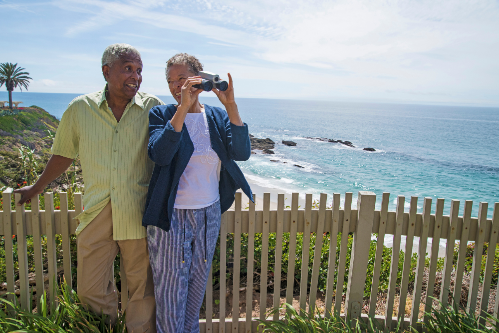 Senior couple outdoors by the ocean, with the woman looking through binoculars.