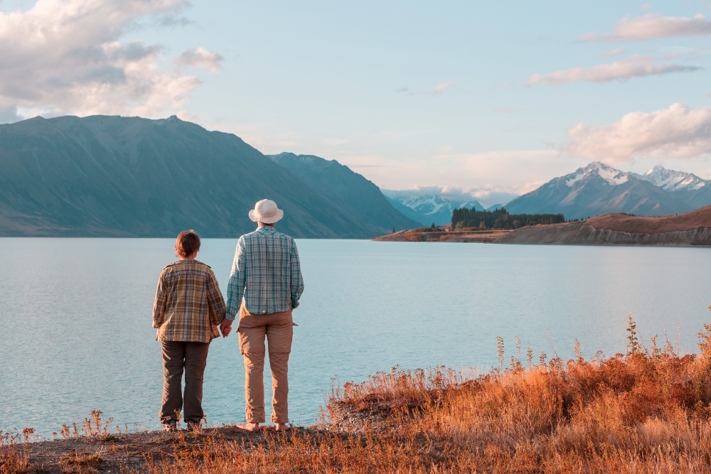 Two people holding hands and looking at a serene lake with mountains in the background.