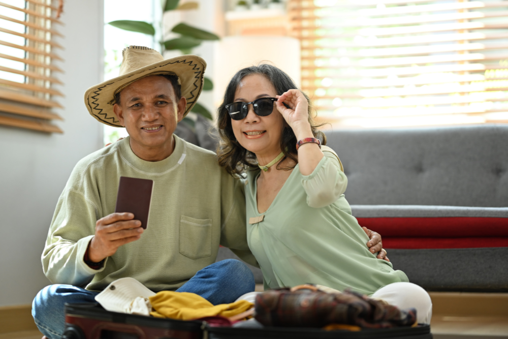 A smiling older couple, with the man wearing a cowboy hat and holding a passport, and the woman wearing sunglasses, sitting next to an open suitcase.