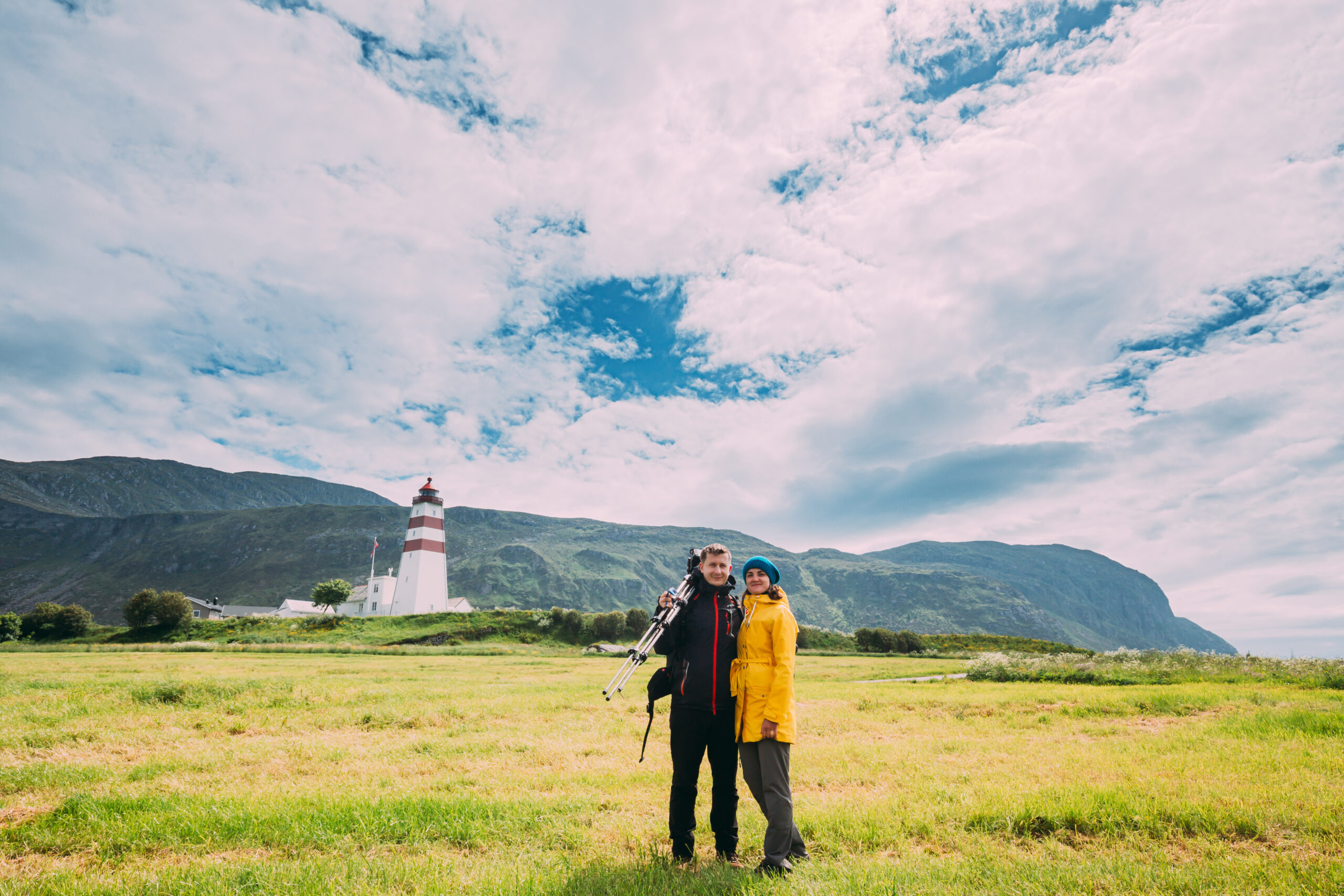 Man and woman standing together in front of a lighthouse with mountains in the background.