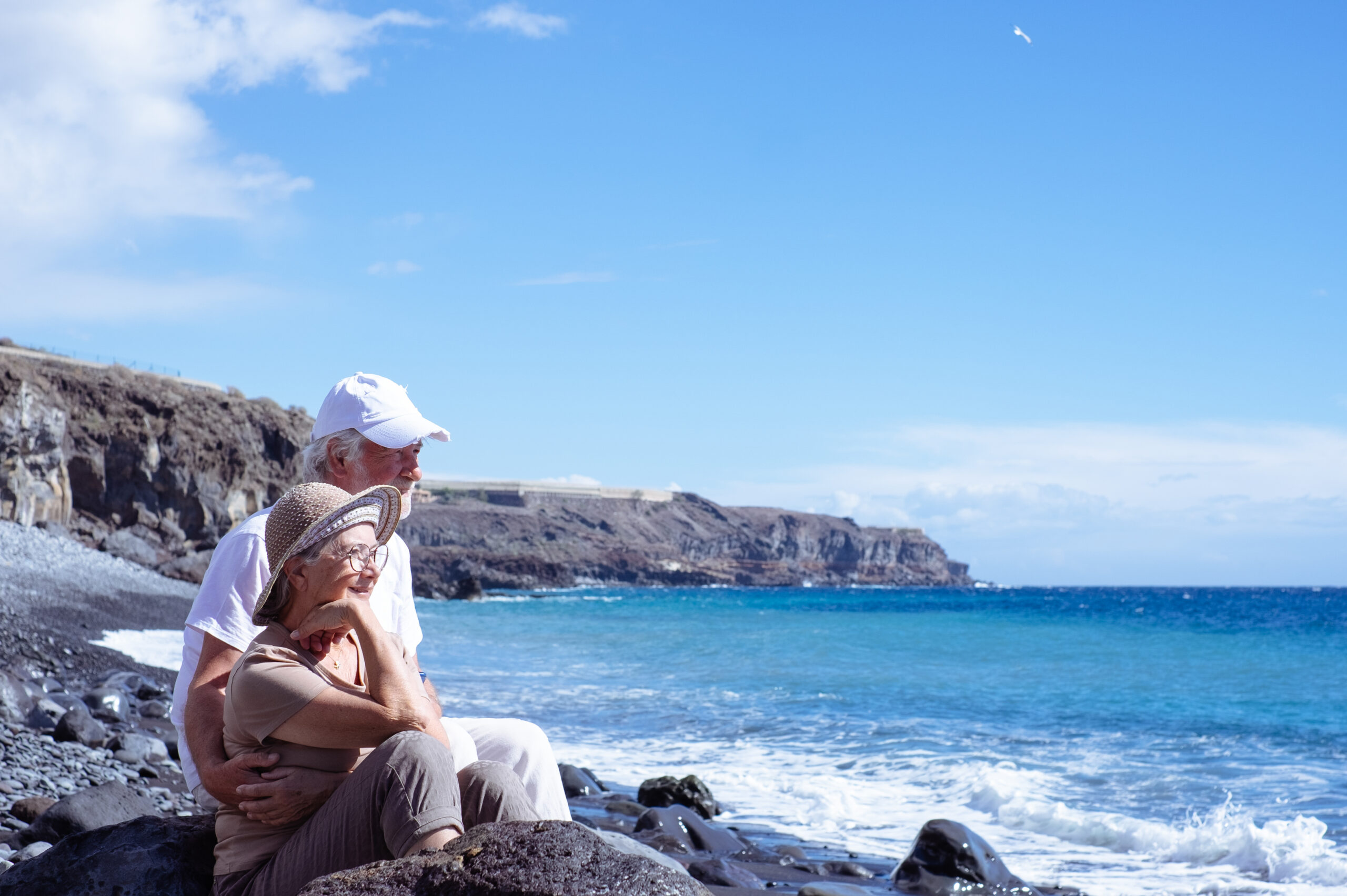 Senior couple sitting on rocks by the ocean, enjoying the view.