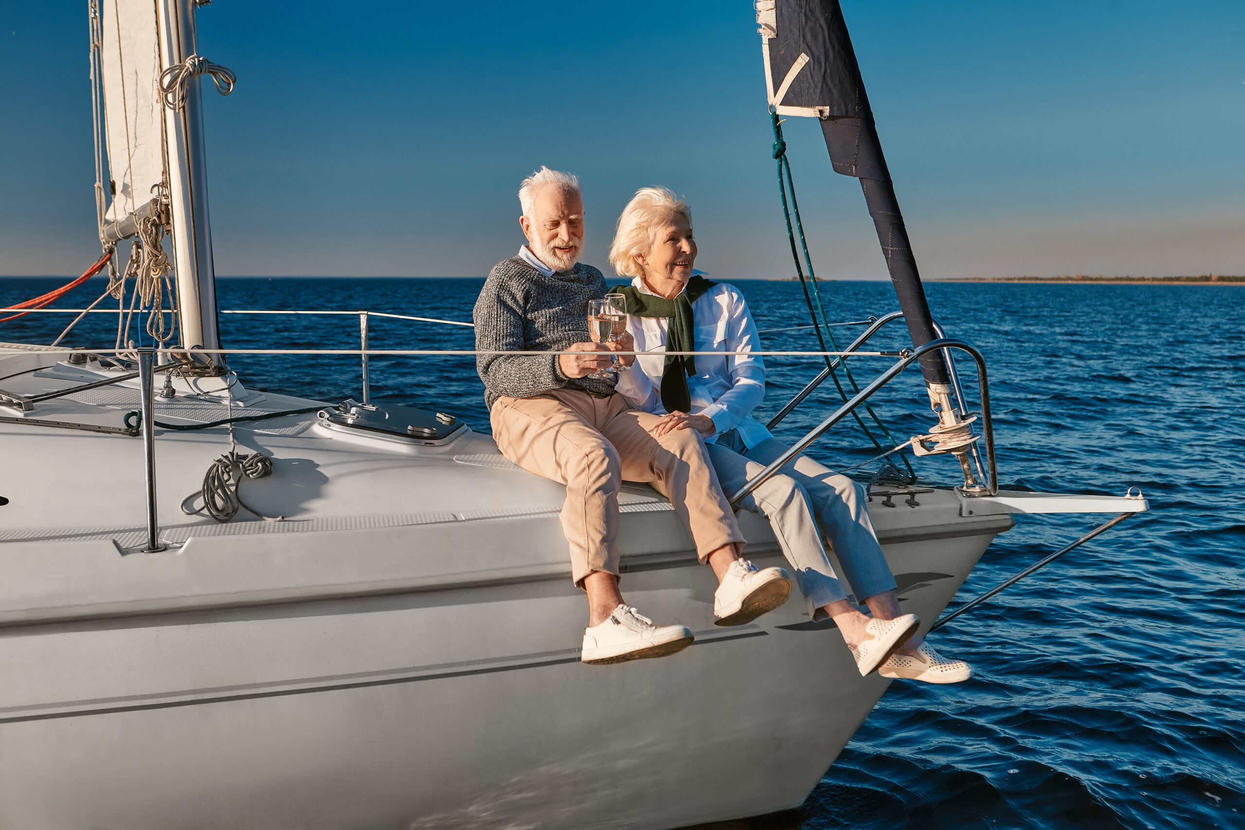 Senior couple sitting on the bow of a sailboat, enjoying a glass of wine together on the open sea.