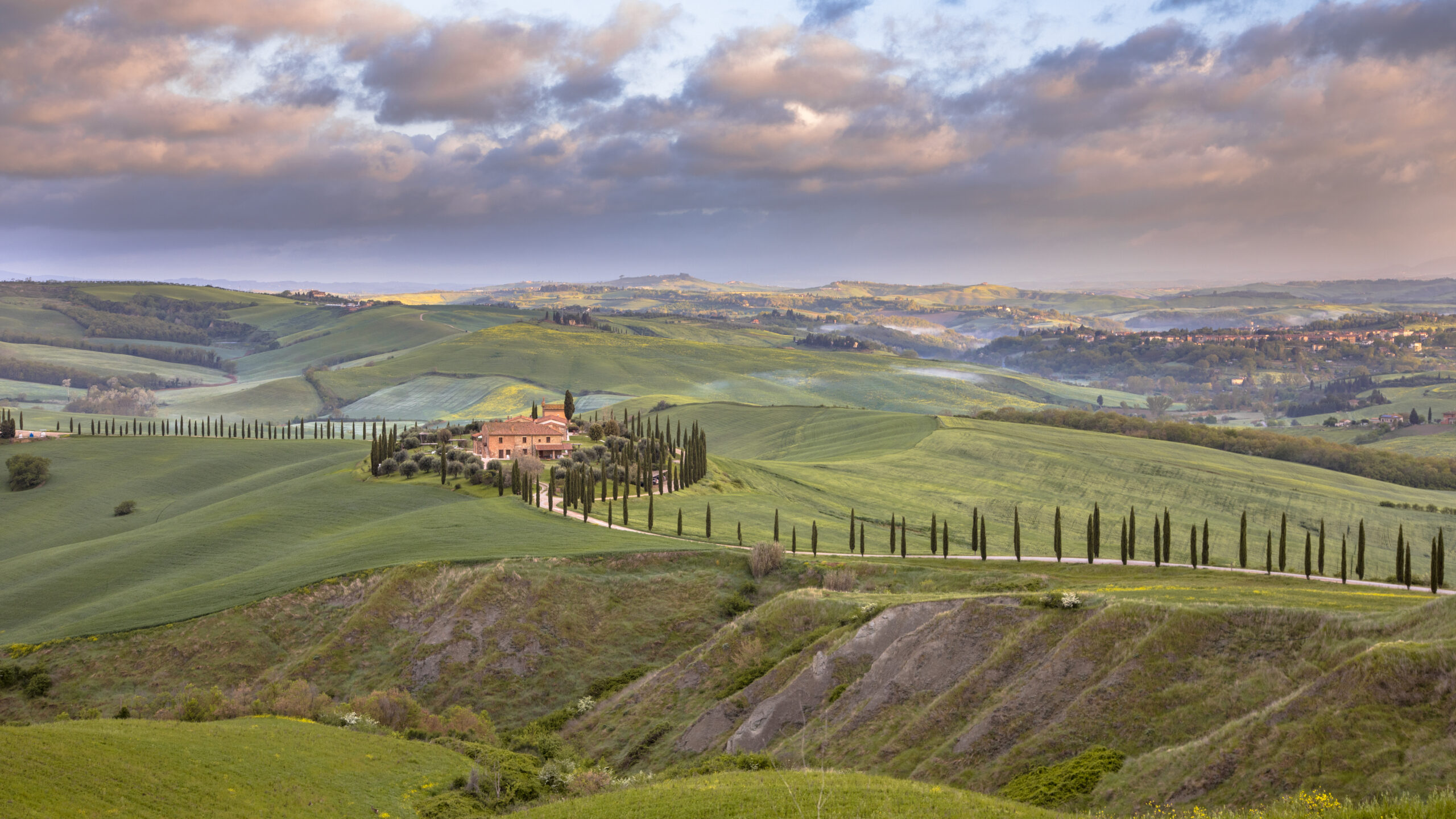 Rolling hills of Tuscany with a farmhouse surrounded by cypress trees at sunset.