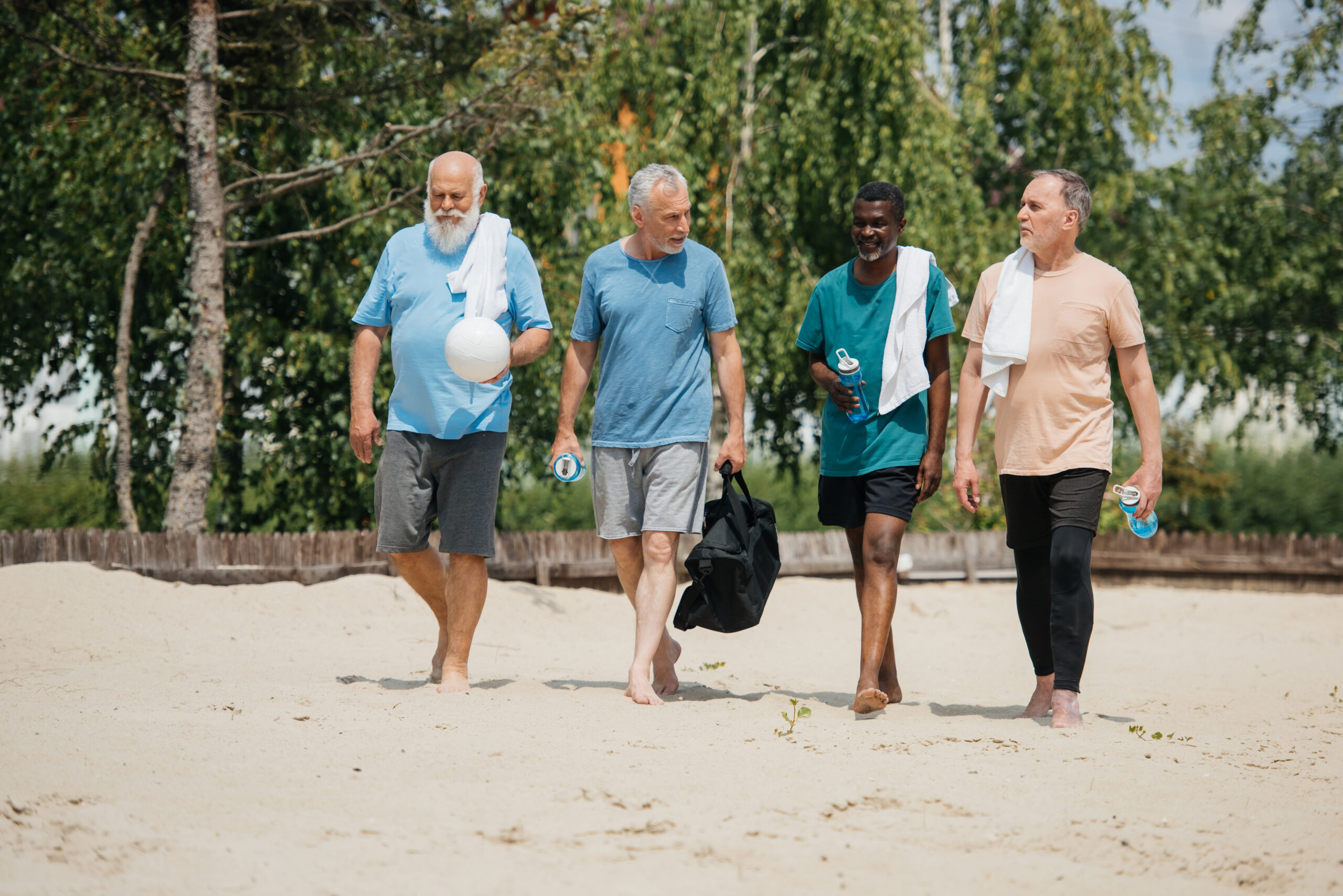 Group of older men walking together on a sandy path, carrying volleyball gear and water bottles.