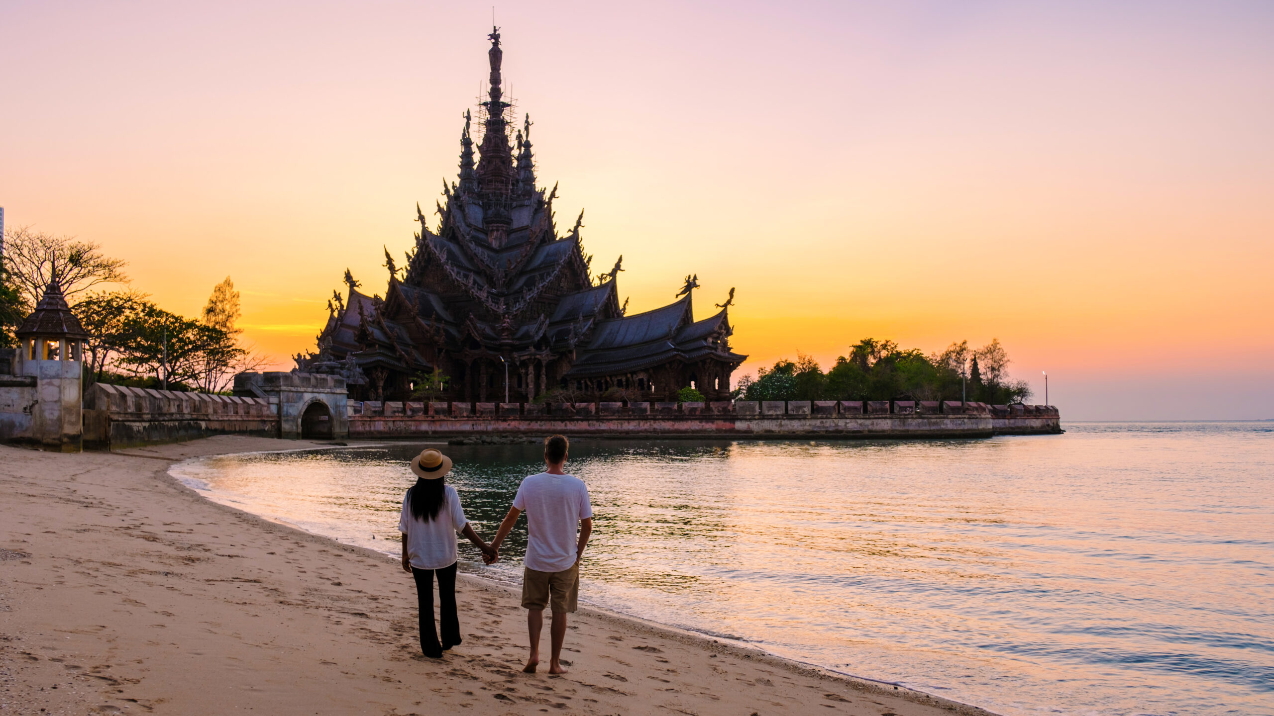 A couple holding hands and walking along the beach towards the Sanctuary of Truth in Pattaya, Thailand, during a stunning sunset.